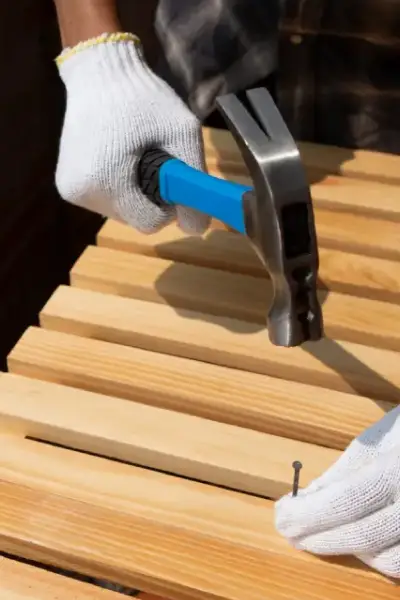 A person wearing gloves hammers a nail into wood slats on a sunny day, focusing intently on the task