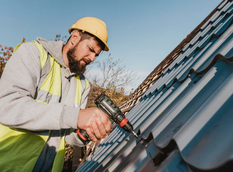 A worker in a yellow hard hat and safety vest is using a power drill to secure metal roofing panels under a clear blue sky